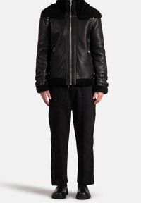 Persona che indossa una giacca in pelle nera con texture e colletto e polsini in shearling, pantaloni neri con dettagli a bottoni e stivali neri.