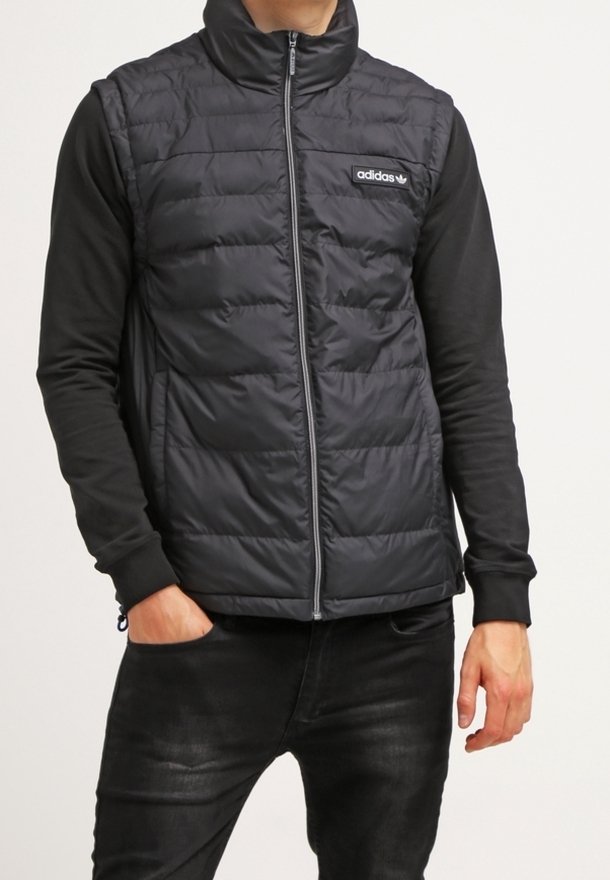 Svart vadderad väst med hög krage, dragkedjestängning och adidas-logotyp på bröstet, buren över en långärmad svart skjorta.