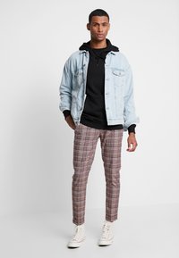 Giacca di jeans blu chiaro su una felpa con cappuccio nera, abbinata a pantaloni a quadretti marroni e sneakers bianche alte.