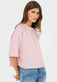 Kvinde med langt brunt hår iført en løs lyserød t-shirt med korte ærmer og blå jeans, stående mod en ensfarvet lys baggrund.