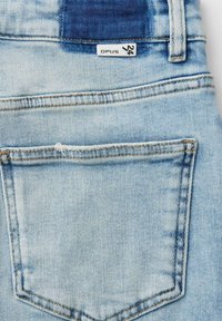 Light blue denim jeans back pocket with visible stitching and a small white fabric tag labeled "Opus 24" near the waistband.