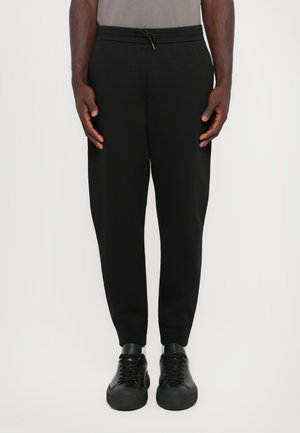 Sorte joggebukser med snor i midjen, ribbestrikkede mansjetter og en glatt tekstur. Båret med sorte joggesko som har plattformsåle.