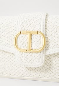 Pochette tissée blanche avec un tissu texturé, fermoir doré avec un design en "T" et une bordure blanche le long des bords.
