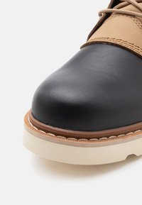 Prim-plan al vârfului unei cizme cu față din piele neagră, partea superioară din piele bej, talpă groasă albă și cusături detaliate de-a lungul marginii.