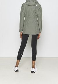 Olivgrüne wasserdichte Jacke mit taillierter Passform, Kapuze und langen Ärmeln, kombiniert mit schwarzen Leggings mit reflektierenden Akzenten und weißen Turnschuhen.