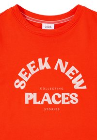 Sweat-shirt à col rond orange vif avec un texte blanc indiquant "CHERCHEZ DE NOUVEAUX ENDROITS" en grandes lettres et "COLLECTER DES HISTOIRES" en lettres plus petites en dessous.