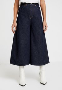 Tummansinisiä farkkuhousuja, jotka ovat löysät ja joissa on etutaskut sekä kontrastivärinen ompelu. Yhdistetty polven yli ulottuviin valkoisiin saappaisiin.