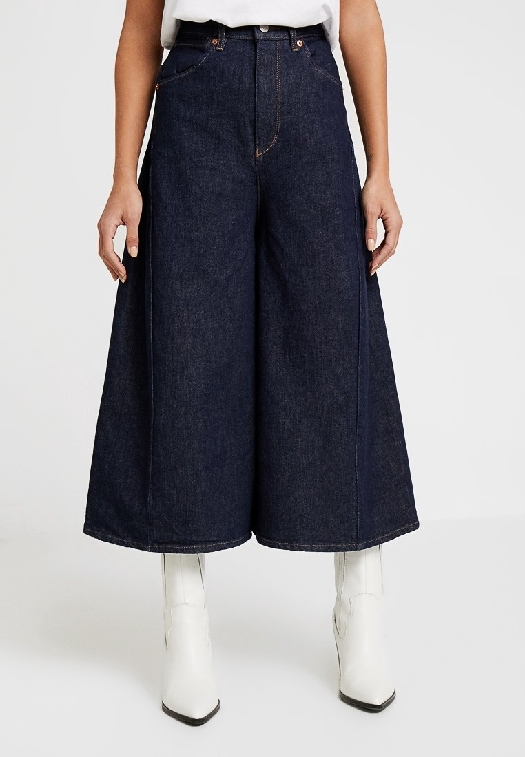 Tummansinisiä farkkuhousuja, jotka ovat löysät ja joissa on etutaskut sekä kontrastivärinen ompelu. Yhdistetty polven yli ulottuviin valkoisiin saappaisiin.