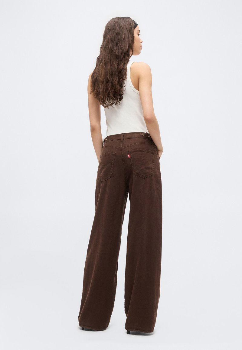 Woman with long wavy hair wearing a sleeveless white top, brown wide-leg pants, and a black headband, standing facing sideways.