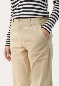 Osoba v béžových kalhotách, s rukou v kapse, a s dlouhým rukávem černobíle pruhovaného trička, pohled od pasu po střed stehno.