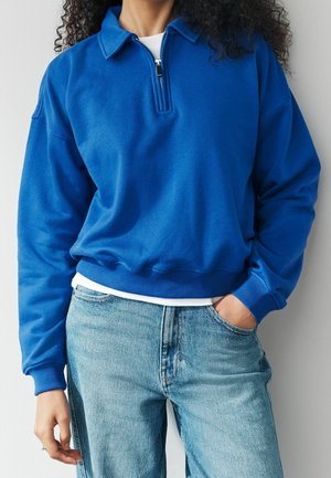Personne portant un pull polaire bleu avec fermeture éclair sur une chemise blanche et un jean bleu clair, une main dans la poche, debout devant un fond uni.