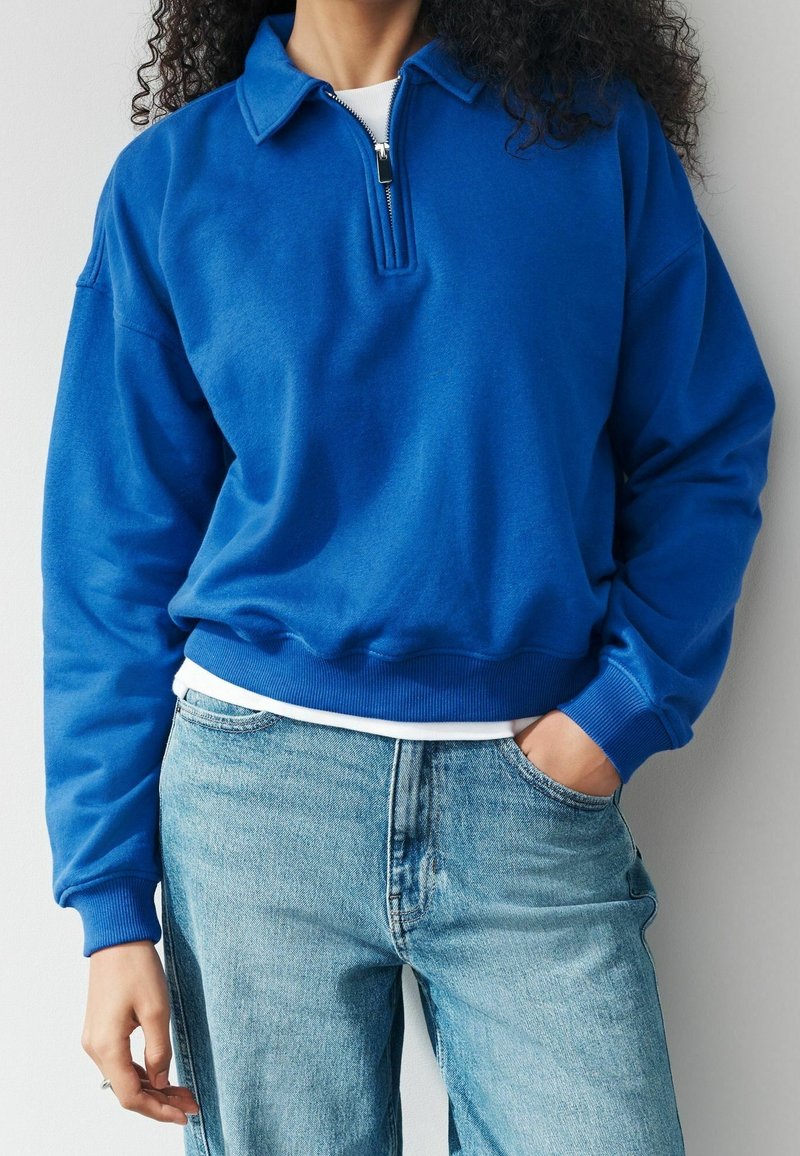 Personne portant un pull polaire bleu avec fermeture éclair sur une chemise blanche et un jean bleu clair, une main dans la poche, debout devant un fond uni.