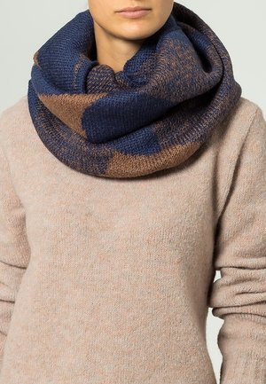 Kobieta nosząca gruby, dziergany niebiesko-brązowy szal typu infinity na beżowym swetrze, zdjęcie od nosa do połowy tułowia.