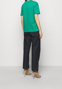 Person som bär en grön kortärmad skjorta, mörka vida jeans och beige högklackade sandaler, stående mot en enkel bakgrund.