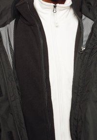 Veste noire avec doublure en mesh, incluant une fermeture éclair à l'avant. En dessous, une couche en polaire blanche avec une fermeture éclair centrale. Tissu texturé visible.
