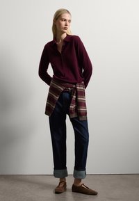 Maglione bordeaux con bottoni abbinato a jeans blu scuro con risvolti. Sciarpa a righe legata in vita. Scarpe marroni dal design minimale.