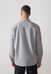 Maglietta grigia a maniche lunghe con colletto a punta, polsini con bottoni e una texture liscia. Presenta uno yoke posteriore con lieve drappeggio.