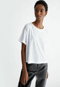 T-shirt bianco a maniche corte con scollatura rotonda, caratterizzato da decorazioni in borchie metalliche disposte in un motivo a griglia sulla parte frontale.