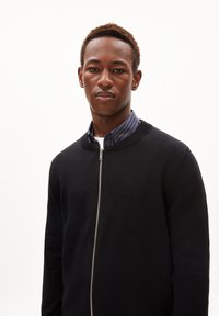 Zwarte ritsvest met een gestructureerde breisel, voorzien van een ronde hals en een contrasterend blauw-wit gestreept shirt eronder.