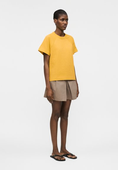 Une femme se tient debout portant un haut jaune moutarde à manches courtes, une jupe plissée beige et des tongs noires devant un fond blanc uni.
