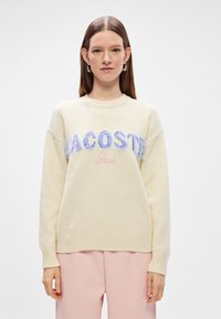 Κρέμα πλεκτή μπλούζα με μπλε λογότυπο "LACOSTE" και ροζ λεπτομέρεια "Παρίσι". Διαθέτει στρογγυλή λαιμόκοψη και ριμπ φινίρισμα στις μανσέτες και στη βάση.