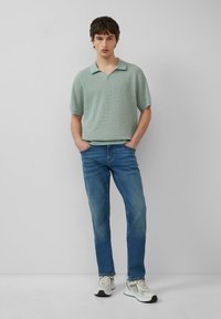 Polo en tricot vert clair avec un col en V et des manches courtes, associé à un jean bleu et des baskets blanches avec des accents noirs.