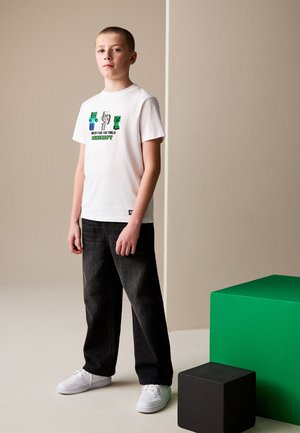 Poika, jolla on valkoinen Minecraft-paita, mustat farkut ja valkoiset lenkkarit, seisoo vihreiden ja mustien kuutioiden vieressä neutraalissa huoneessa.