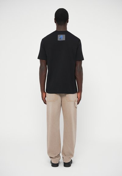 T-shirt en coton noir à manches courtes, avec un patch logo bleu dans le dos. Porté avec un pantalon beige ample et des chaussures noires.