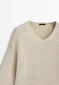 Pull tricoté de couleur crème avec un tissage lâche et un col en V, posé à plat sur une surface blanche.