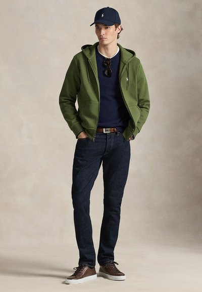 Πράσινο φούτερ με φερμουάρ, μπλε πουλόβερ, σκούρο τζιν, καφέ αθλητικά παπούτσια και μπλε καπέλο. Γυαλιά ηλίου κρέμονται από το λαιμό. Απλός σχεδιασμός, casual στυλ.