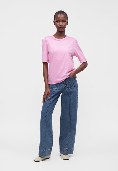 Femme debout, une main dans la poche, portant un haut rose clair à manches courtes, un jean bleu à jambes larges et des chaussures crème sur un fond uni.