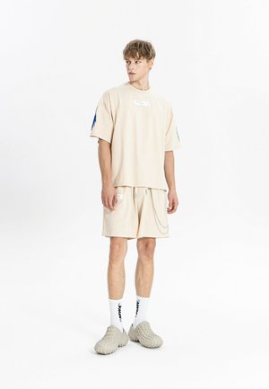 Joven de pie, lleva una camiseta beige holgada y pantalones cortos con detalle de cadena, calcetines blancos con texto y zapatos beige texturizados sin cordones.