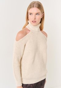 Pull en tricot côtelé beige avec un col haut, des épaules dénudées et des manches longues. La texture semble douce et épaisse.
