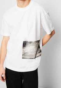 Bílé bavlněné tričko s obdélníkovým potiskem s černým textem "Calvin Klein" a "Calvin Klein Jeans" na spodní přední straně.