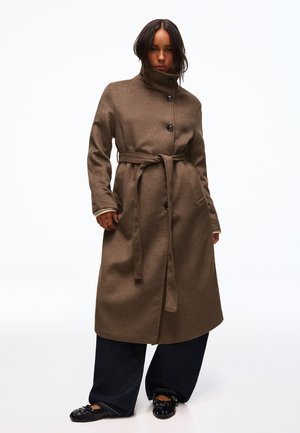 Femme portant un long manteau marron ceinturé avec un col montant, un pantalon noir à jambes larges et des chaussures plates noires, debout sur un fond blanc.