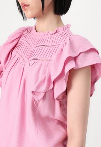 Blouse rose en tissu léger, avec des manches courtes volantées et un corsage texturé aux accents de couture décoratifs.