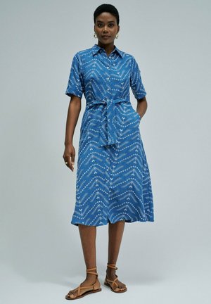 Mujer con vestido midi azul con estampado, mangas cortas, botones delanteros, cinturón en la cintura y sandalias marrones con pulsera en el tobillo, de pie frente a un fondo liso.