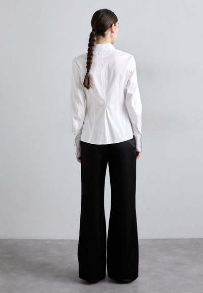 Camicia bianca aderente con maniche lunghe, polsini abbottonati e colletto alto, abbinata a pantaloni neri a gamba larga, mostrata da dietro.