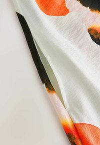 Tissu blanc avec un grand imprimé floral orange, noir et rouge et une fente latérale sur un fond clair.