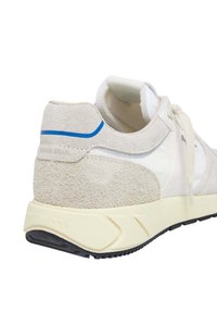 La zapatilla presenta una parte superior de ante color beige claro con un panel de tela blanca, una línea de acento azul y una suela intermedia texturizada de color crema con suela exterior negra.