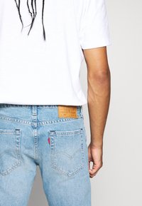 Ljusblå denimjeans med blekt utseende, med en brun läderlapp på baksidan, fem fickor och röd logodetalj på bakfickan.