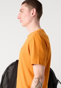 Orange bomulds t-shirt med korte ærmer, rund hals og et lille logo foran, båret sammen med en sort rygsæk på modellens skulder.