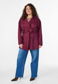 Bordeauxroter Doppelreiher-Mantel mit Bindegürtel, weiten Ärmeln und zwei Knöpfen; kombiniert mit weit geschnittenen blauen Jeans und braunen spitzen Absätzen.