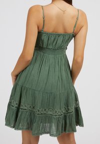 Vestido verde sin mangas con bodice fruncido y falda de capas con volantes. Presenta acentos de encaje y una tela ligera y texturizada.