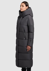 Lungo cappotto nero voluminoso con colletto alto, caratterizzato da sezioni orizzontali trapuntate e una tasca laterale con cerniera. Texture liscia e morbida.