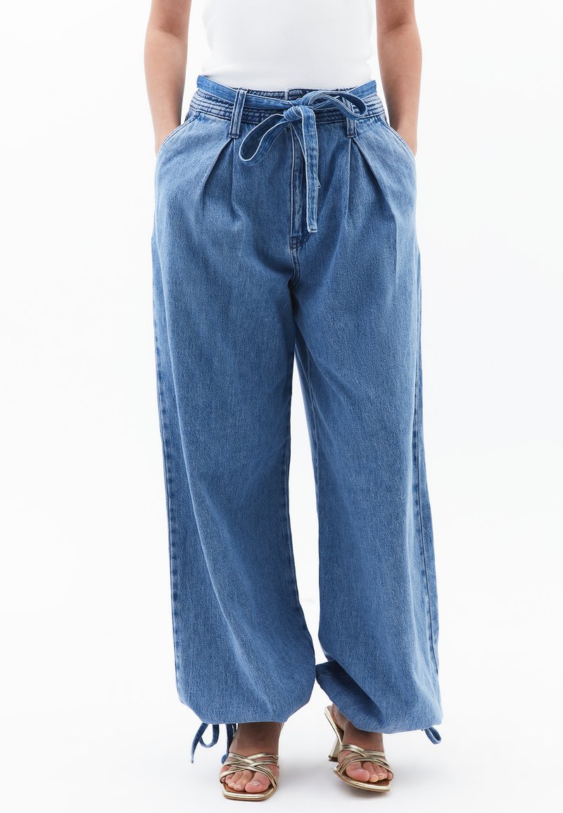 Pantalones de mezclilla anchos en azul claro, con cintura ajustable mediante cordón y plisados. Presentan lazos en los tobillos y un corte relajado. Tejido texturizado con un acabado suave.