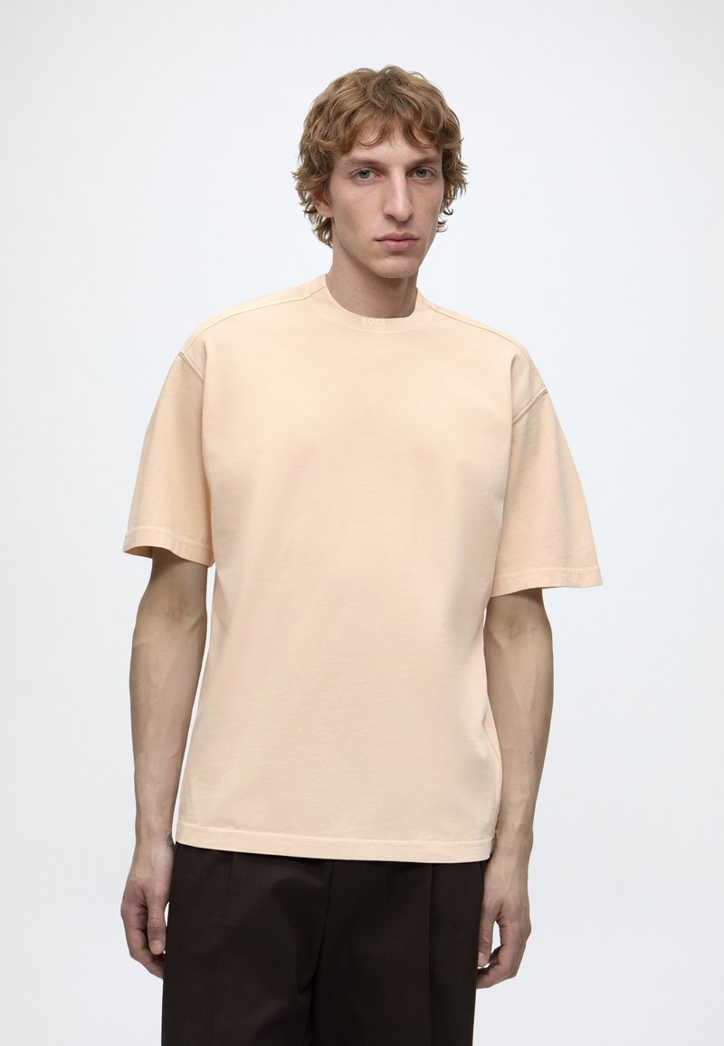 Jeune homme aux cheveux bouclés châtain clair portant un t-shirt beige à manches courtes et un pantalon foncé, debout devant un fond blanc uni.