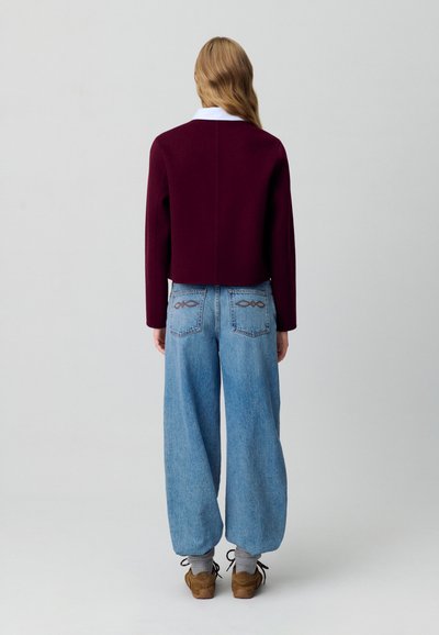 Suéter burdeos corto de mangas largas, combinado con jeans claros sueltos que presentan costuras decorativas en los bolsillos traseros. Zapatos marrones visibles.