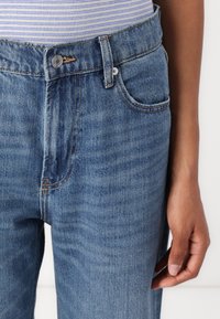 Jeans de ganga azul clara com um botão na cintura, bolsos frontais e costura em contraste. O tecido tem uma textura suave e um acabamento ligeiramente desbotado.