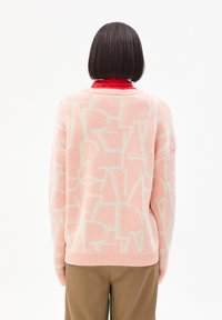 Una persona con capelli corti e scuri indossa un maglione rosa a fantasia sopra una camicia rossa e pantaloni marroni, rivolta verso l'esterno su uno sfondo bianco semplice.